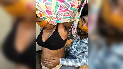 Media: Video of two African women in a healthcare setting. The woman on the left wears a colorful headscarf, black bra, and beige pants; the woman on the right, wearing a plaid shirt, gently holds her abdomen.