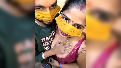 Media: Video: A couple in traditional attire, wearing yellow masks, with a woman in a pink blouse and the man in a green shirt, touching each other's faces affectionately.