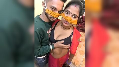 Media: Video of a man and woman in a home setting. The man, wearing a green sweatshirt, has a yellow mask over his eyes. The woman, in a red sari, has a yellow mask over her eyes.