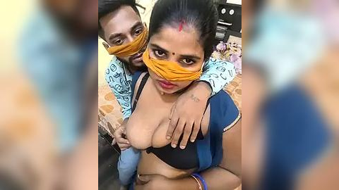 Media: A video of a South Asian woman with medium brown skin and a bindi, wearing a yellow mask, black bra, and blue skirt, sitting on a bed with a man behind her, both looking at the camera.