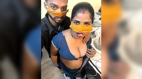 Media: Video of two South Asian women in masks, one with a blue blouse exposing cleavage, standing close together. Background includes a blurred figure and a bed with a fan.