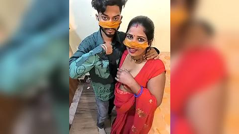 Media: Video of a smiling South Asian couple, both wearing orange masks, in traditional attire, standing on a wooden floor in a home.