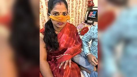 Media: Video of a woman in a red sari with a yellow mask, sitting on a bed with a child in blue clothing, in a room with patterned curtains.