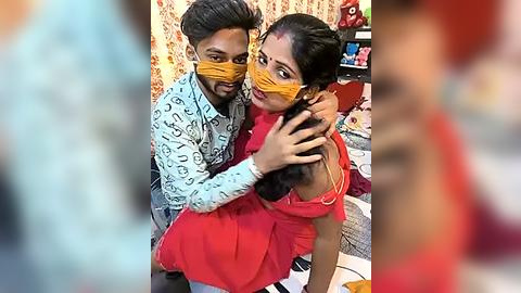 Media: A video of a young South Asian couple wearing yellow masks, the man in a light blue patterned shirt, the woman in a red dress, hugging in a cluttered, colorful room with floral wallpaper and a toy shelf.