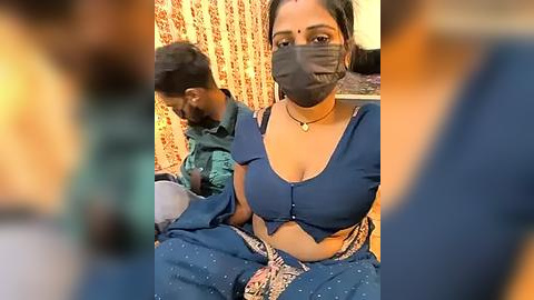 Media: Video of two South Asian women wearing blue sarees and face masks, sitting closely together, background with ornate wallpaper.