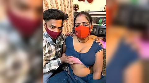 Media: Video of a South Asian couple, with a man in a plaid shirt and a woman in a blue crop top, sitting on a sofa, both wearing red face masks, in a cozy, dimly lit room with floral curtains.