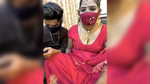 Media: Video of a woman in a red sari with gold embroidery, a face mask, and gold jewelry, sitting next to a young boy in a black shirt.