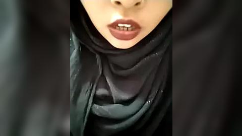 Media: A close-up video of a person's face, wearing a black hijab and maroon lipstick, with visible teeth. The background is blurred, emphasizing the subject's face.