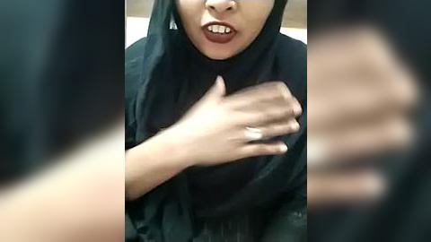 Media: Video of a young woman with light brown skin, wearing a black hijab, mouth open in a wide, exaggerated smile, hands clasped in front, wearing a silver ring.