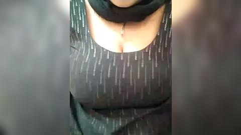 Media: Video of a woman's torso in a black, textured dress with vertical white lines, showcasing cleavage. Her skin is light, and she wears a delicate necklace. The background is blurred.