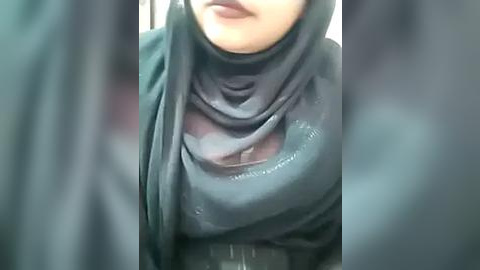 Media: A video of a woman wearing a black hijab, partially revealing her chest area with visible nipples through the sheer fabric, standing indoors with blurred background.