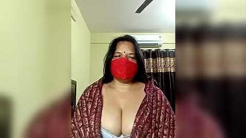 Media: A video of an Indian woman with a medium complexion, wearing a red mask, a maroon patterned robe, and a white lace bra, in a dimly lit room with yellow walls and a brown curtain.