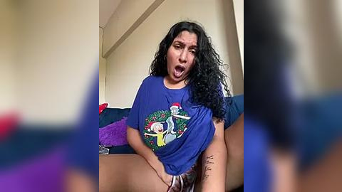 Media: Video of a woman with long black hair, wearing a blue T-shirt, sitting on a couch, mouth open in surprise. Background shows a beige wall and purple cushions.