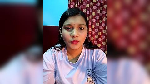 Media: Video of a young South Asian woman with medium brown skin, dark hair, and red lipstick, wearing a white shirt with a cartoon character on the left breast. Background includes red curtains with a floral pattern and a light blue wall.