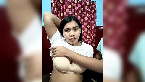 Media: Video of a young woman with medium brown skin, dark hair, and medium build, wearing a beige bra, lifting her white T-shirt to expose her chest. Background features a red curtain with floral patterns and a blue wall.