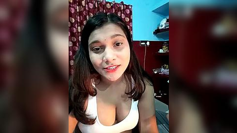 Media: Video of a young South Asian woman with medium skin tone, dark hair, and a slim build, smiling in a white tank top with large cleavage, sitting in a room with red curtains and blue walls.