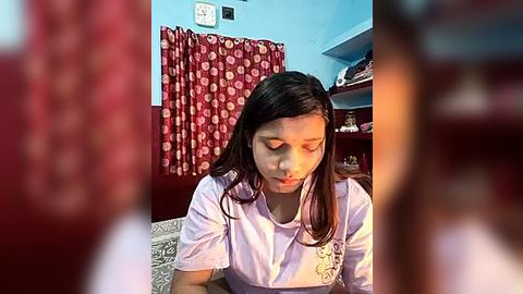 Media: A video of a young woman with long dark hair, wearing a light pink shirt, seated indoors. Red curtains with white polka dots hang behind her, and shelves with personal items are visible in the background.