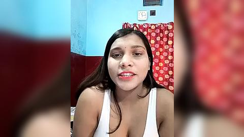 Media: Video of a young South Asian woman with medium skin tone, dark hair, wearing a white tank top, smiling, in a bathroom with blue walls, red curtains, and visible electrical outlets.