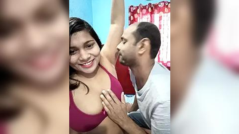 Media: Video of a South Asian woman with medium skin tone and brown hair, smiling, in a magenta bra, and a bald man in a white shirt, kissing her chest in a bedroom with blue walls and red curtains.