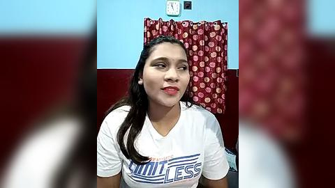 Media: Video of a young woman with light skin, long dark hair, and a white t-shirt with \"Billionaire\" in blue text. She's indoors with a red-patterned curtain and a clock on the wall in the background.