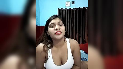 Media: A video of a young South Asian woman with medium skin tone, wearing a white tank top, smiling with closed eyes. She is in a cozy room with blue walls, dark curtains, and a red couch.