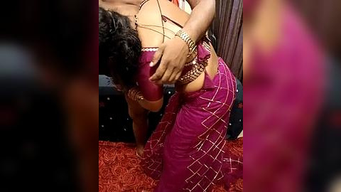 Media: Video of a South Asian woman in a pink saree, leaning forward on a red-patterned carpet, with a man's hands on her hips, in a dimly lit room.