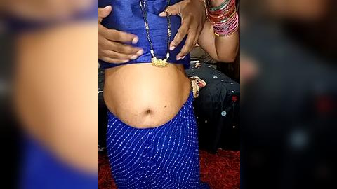 Media: A video of a pregnant woman in a blue polka-dot sari, revealing her belly, with hands on her stomach, wearing a gold pendant. Background shows a blurred red carpet and colorful bangles.