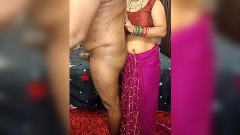 Media: Video of a nude man standing next to a woman in a vibrant pink saree with a black blouse, wearing colorful bangles. Background shows a bed with red and black bedding.