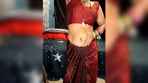 Media: Video of a woman wearing a red saree with a gold necklace, bangles, and a beaded bracelet, standing against a blue and red cushioned backdrop.