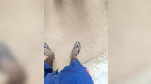 Media: A video captures a pair of bare feet in blue sandals and red shorts standing on a dusty, sandy floor. The perspective is from above, emphasizing the texture and contrast of the ground.