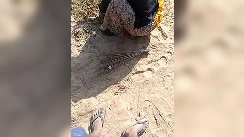 Media: Video of a person's feet wearing sandals on a dusty, barren ground. The person is seated, wearing a colorful skirt and a black shirt. Shadows suggest a sunny day.