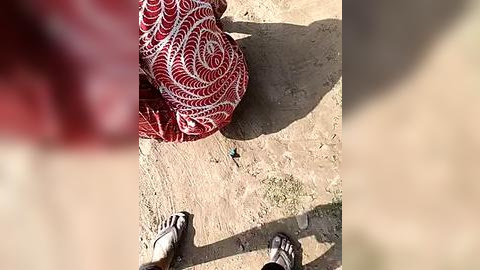 Media: Video of a person sitting on the ground with a red and white patterned cloth, wearing dark sandals and a red shirt, surrounded by dry, sandy terrain.