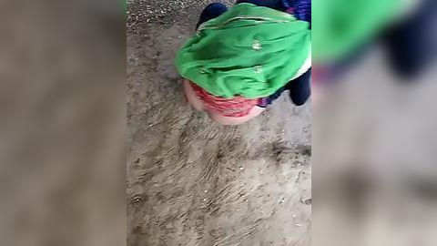 Media: Video of a child wearing a green shirt and red shorts, standing on a sandy ground, with blurred background.