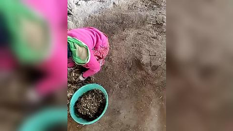 Media: A video of a person, wearing a bright pink jacket and green pants, kneeling on dirt, scooping soil with a teal bowl into a larger green container. The background is a blurred, earthy brown.