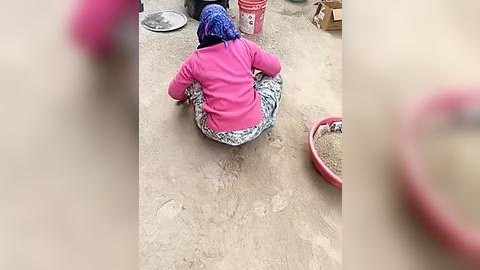 Media: Video of a woman with a blue headscarf and pink jacket, sitting on a patterned cushion, sorting rice on a dirt floor.