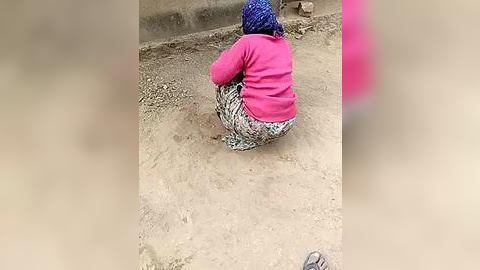 Media: Video of an elderly woman, seated on the ground, wearing a bright pink sweater, patterned skirt, and blue knitted hat, against a dusty, bare background.