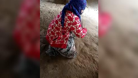 Media: Video of a person kneeling in a dirt floor room, wearing a red and white patterned jacket, blue headscarf, and patterned pants.