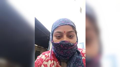 Media: Video of a woman with light brown skin, wearing a blue and purple floral-patterned hijab and red bindi, standing indoors with a blurred background.