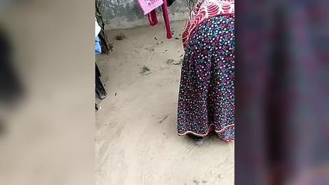 Media: Video of a woman in a colorful floral dress standing on a dirt path. A pink plastic chair is partially visible behind her. The background shows a rough, gray wall and scattered vegetation.