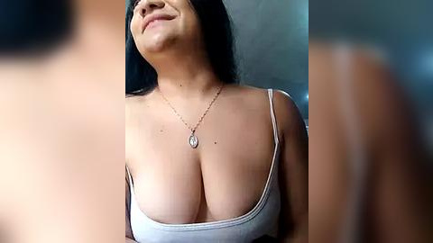 Media: Video of a smiling, light-skinned woman with long black hair, wearing a low-cut gray tank top revealing ample cleavage, and a silver necklace. Out-of-focus figures in the foreground.