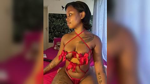 Media: Video of a dark-skinned woman with short hair, wearing a red, crisscross lingerie set with large bow details, sitting on a bed with pink sheets. Her tattoos are visible. Background includes a black and white wall and a window with sheer curtains.