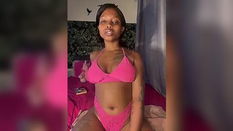 Media: A video of a slender Black woman with medium-length, straight hair, wearing a pink lace lingerie set, sitting on a bed with a pink blanket, in a dimly lit room with a dark green wall and a butterfly sticker.