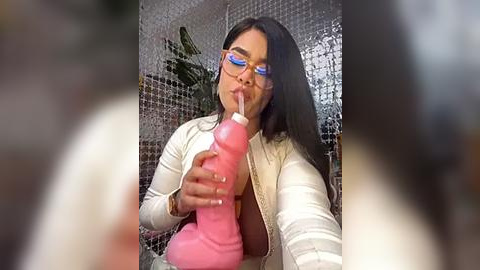 Media: Video of a woman with long dark hair, wearing glasses and a white cardigan, holding a pink dildo to her mouth in a bathroom with a tiled wall and green plant.