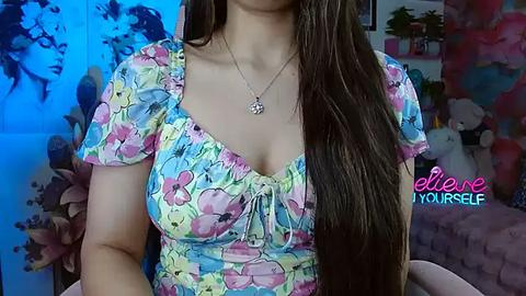 Media: Video of a woman with long brown hair, wearing a colorful floral top, necklace, and a blue background.