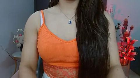 Media: Video of a woman with long brown hair, wearing a bright orange tank top with lace trim, and a silver necklace, seated indoors with a pink floral arrangement and decorative ornaments in the background.