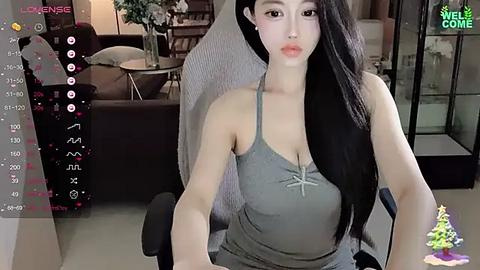 Media: Video of an Asian woman with long black hair in a gray tank top, sitting on a black chair in a modern living room.