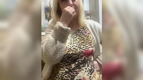 Media: Video of an older, fair-skinned woman with shoulder-length blonde hair, wearing a leopard-print dress and beige cardigan, covering her mouth in a gesture of surprise or shock.