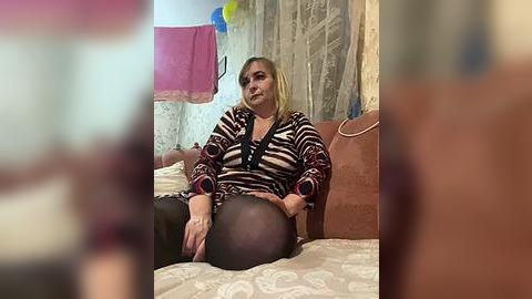 Media: A video of a Caucasian woman with blonde hair, wearing a zebra-patterned top and black tights, sitting on a beige bed in a room with a pink towel hanging on a wall.