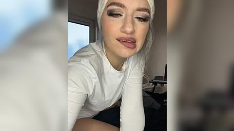 Media: Video of a young Caucasian woman with fair skin, light blonde hair, and dark eyebrows, wearing white makeup and a white top, leaning forward, in a modern indoor setting.