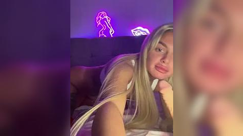 Media: Video of a blonde woman lying on a bed, partially blurred in the foreground, wearing a white bra. Neon purple light illuminates the background, featuring a silhouette.